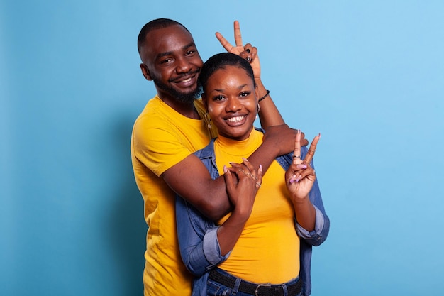 Photo gratuite couple étreignant et montrant un signe de paix avec les doigts devant la caméra, se sentant heureux dans sa relation. portrait d'un homme et d'une femme joyeux dans une étreinte faisant un geste de symbole v avec les mains et souriant.