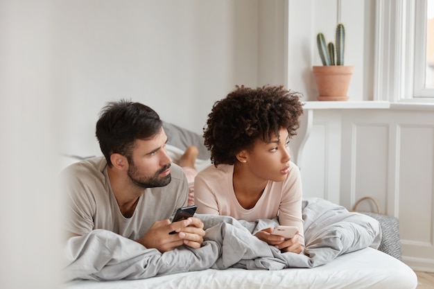 Un couple familial contemplatif utilise des cellules cellulaires, concentré sur le côté, s'allonge au lit le matin