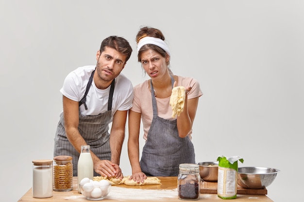 Photo gratuite un couple de famille épuisé et mécontent a des problèmes avec la consistance de la pâte, ne peut pas sculpter le pain, porte des tabliers, passe beaucoup d'heures à la cuisine, a un échec culinaire. les employés du restaurant préparent le gâteau