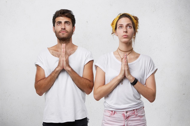 Photo gratuite couple de famille priant ensemble pour la santé de leur enfant. jeune homme barbu et jolie femme en t-shirts blancs gardant leurs mains ensemble tout en priant en levant les yeux pleins d'espoir pour mieux