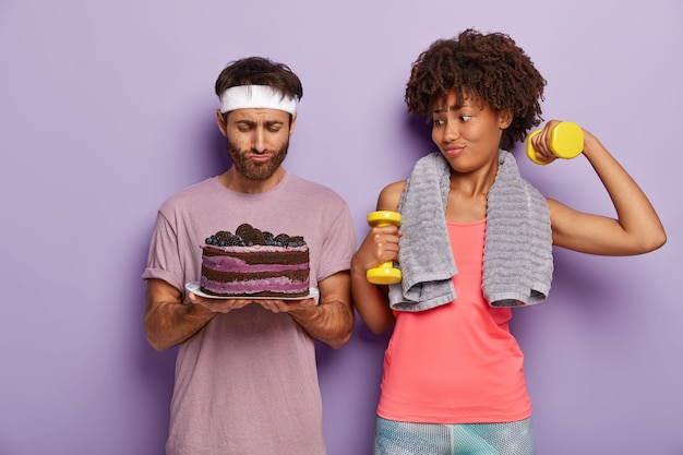 Un couple de famille s'entraîne activement, vêtu de vêtements de sport, soulève des haltères, se tient l'un à côté de l'autre, regarde avec tentation sur le gâteau