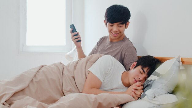 Couple gay asiatique utilisant un téléphone portable à la maison. Un jeune homme LGBTQ + asiatique heureux se détendre après le réveil, vérifiez les médias sociaux pendant que son petit-ami dort allongé sur le lit dans sa chambre à la maison le matin.