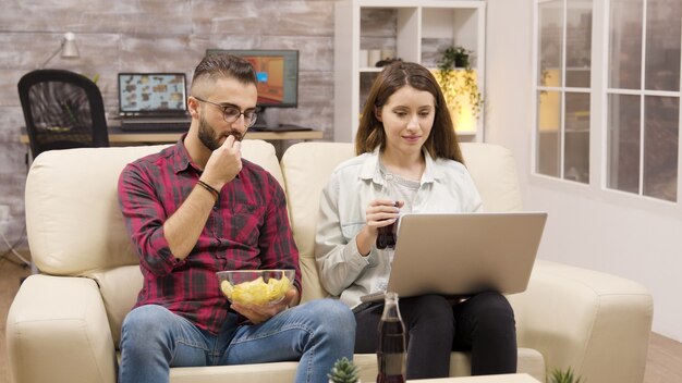 Couple heureux sur le canapé mangeant des chips et regardant un ordinateur portable