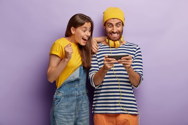 Un couple heureux excité utilise un téléphone portable pour jouer à des jeux en ligne, regarde de manière impressionnante le smartphone, est obsédé par les technologies modernes, habillé de vêtements à la mode. addiction à Internet
