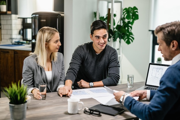 Un couple heureux se tenant la main tout en parlant de plans de logement avec un agent immobilier à la maison