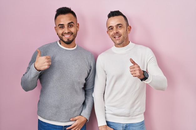 Couple homosexuel debout sur fond rose faisant un geste heureux avec la main approuvant l'expression regardant la caméra montrant le succès