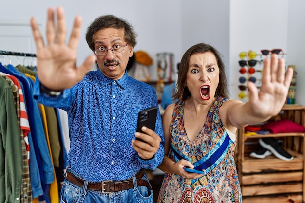 Photo gratuite couple interracial d'âge moyen dans un magasin de détail à l'aide d'un smartphone faisant un geste d'arrêt avec les mains paumes expression de colère et de frustration