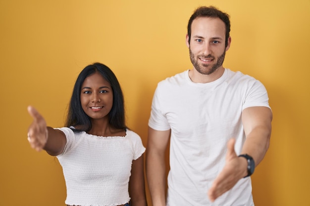 Photo gratuite couple interracial debout sur fond jaune souriant amical offrant une poignée de main comme salutation et accueil. entreprise prospère.