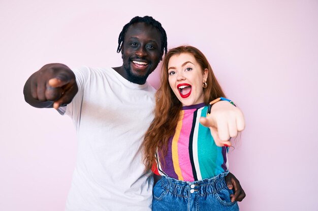 Couple interracial portant des vêtements décontractés pointant vers vous et la caméra avec les doigts, souriant positif et joyeux