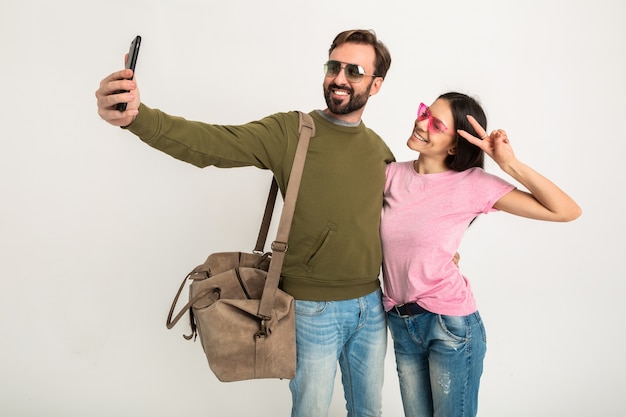 Couple Isolé, Jolie Femme Souriante En T-shirt Rose Et Homme En Sweat-shirt Avec Sac De Voyage, Portant Des Jeans Et Des Lunettes De Soleil, S'amusant, Voyageant Ensemble Faisant Selfie Drôle Photo Sur Téléphone