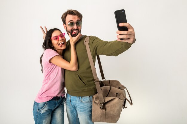 Couple isolé, jolie femme souriante en t-shirt rose et homme en sweat-shirt avec sac de voyage, portant des jeans et des lunettes de soleil, s'amusant, voyageant ensemble faisant selfie drôle photo sur téléphone