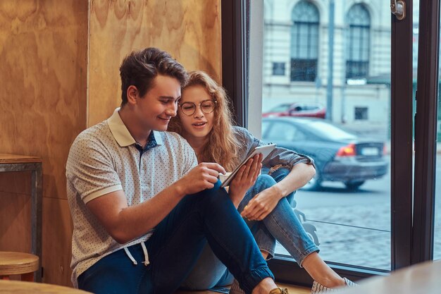 Couple de jeunes étudiants heureux utilisant une tablette numérique assis sur un rebord de fenêtre à la cantine du collège pendant une pause.