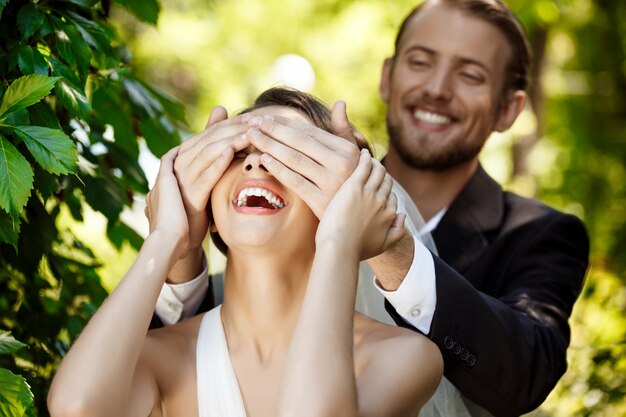 Couple de jeunes mariés souriant. Marié couvrant les yeux de la mariée avec les mains.