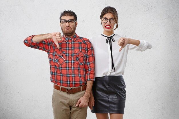 Un couple malheureux de professeurs professionnels montre un signe négatif, garde le pouce baissé, n'est pas d'accord avec quelque chose.