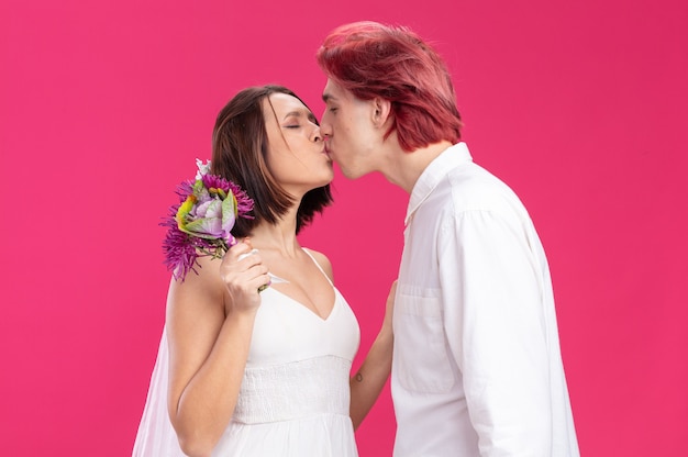 Couple de mariage heureux en robe de mariée avec des fleurs heureux amoureux s'embrasser debout sur un mur rose