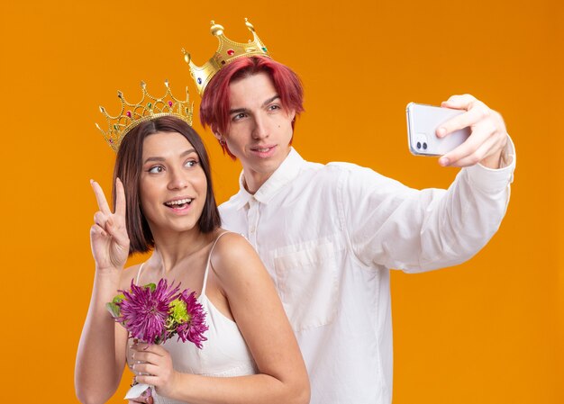 Couple de mariage marié et mariée avec bouquet de fleurs en robe de mariée portant des couronnes d'or souriant joyeusement faisant selfie à l'aide d'un smartphone debout sur un mur orange