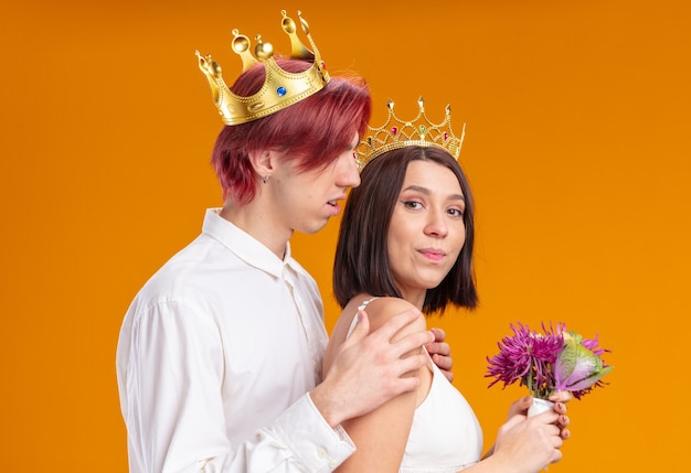 Couple de mariage marié et mariée avec bouquet de fleurs en robe de mariée portant des couronnes d'or souriant joyeusement posant ensemble heureux en amour