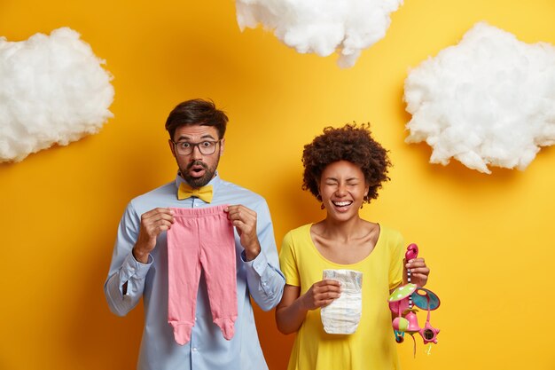 Un couple marié attend un enfant. Mari et femme posent avec des trucs pour bébé, femme enceinte afro-américaine rit joyeusement, tient la couche et mobile, futur père choqué pose avec des vêtements pour nouveau-né