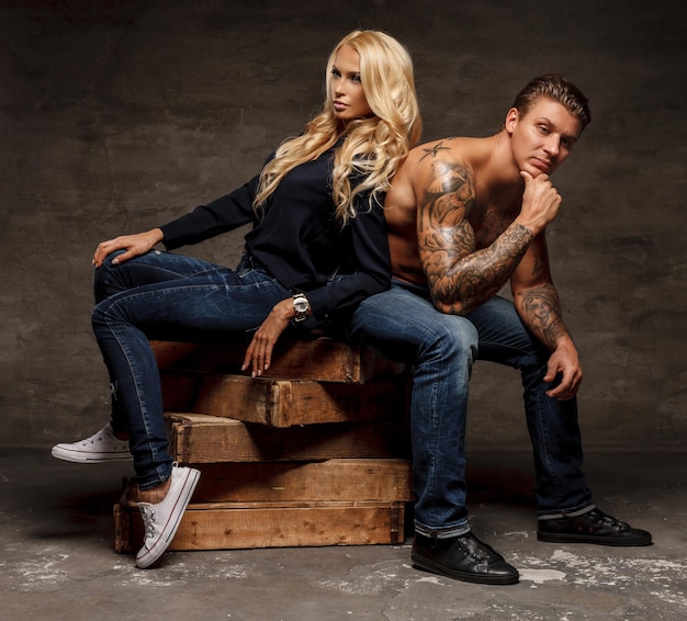 Photo gratuite couple à la mode moderne. homme tatoué torse nu et femme blonde sensuelle assis sur des blocs de bois.