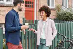 Photo gratuite couple multiracial positif à pied en milieu rural, se promener le week-end, boire du café à emporter, se tenir près d'une clôture, discuter agréablement les uns avec les autres