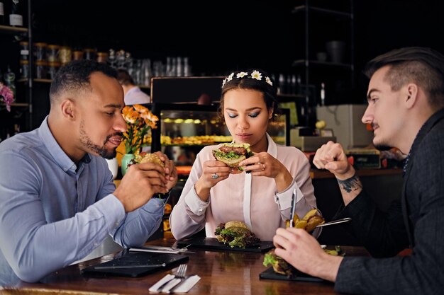 Le couple noir américain et un hipster caucasien mangent des sandwichs végétariens dans un café.