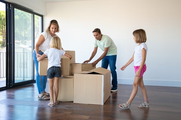 Couple de parents et deux filles ouvrant des boîtes et déballant des choses dans leur nouvel appartement vide