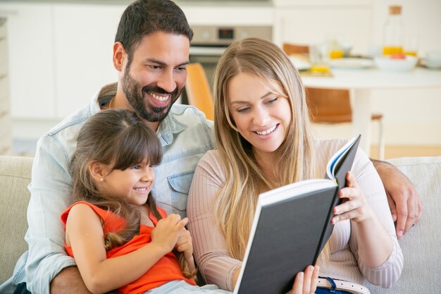 Couple de parents joyeux et petite fille aux cheveux noirs assis sur un canapé dans le salon, lisant un livre ensemble et riant.