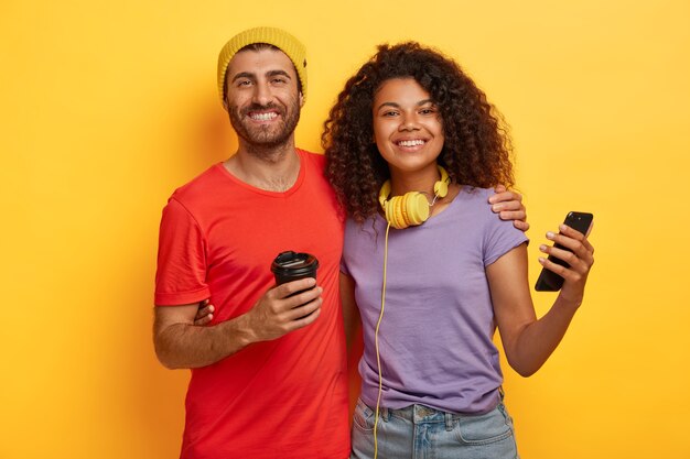 Couple passe du temps libre ensemble, boit du café et utilise un téléphone cellulaire moderne pour la communication en ligne, vêtu de t-shirts, se tient près l'un de l'autre sur fond jaune