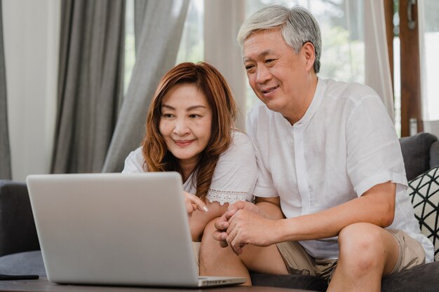 Couple de personnes âgées asiatique utilisant un ordinateur portable à la maison. Grands-parents chinois d'origine asiatique, surfez sur Internet pour consulter les médias sociaux en position couchée sur le canapé dans le concept de salon à la maison.