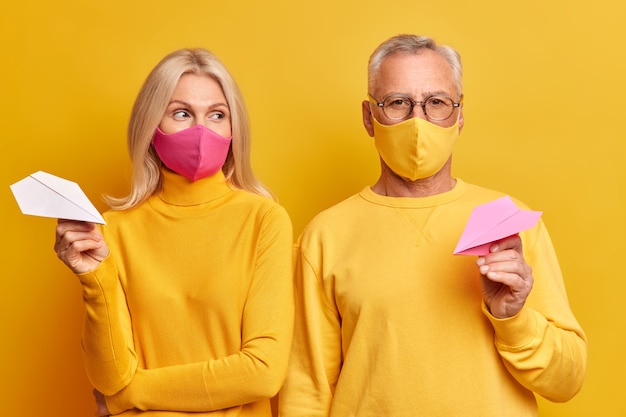 Un couple de personnes âgées porte des masques jetables pour se protéger de la maladie du coronavirus rester à la maison pendant la quarantaine vêtus de vêtements jaunes décontractés tenir des avions en papier faits à la main posent en studio