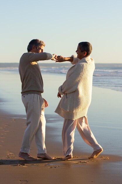 Un couple de personnes âgées romantique passe du temps au bord de la mer au coucher du soleil, s'amuse et danse pieds nus sur du sable mouillé. Dame aux cheveux courts tenant la main de l'homme et riant. Loisirs, romance, concept de relation