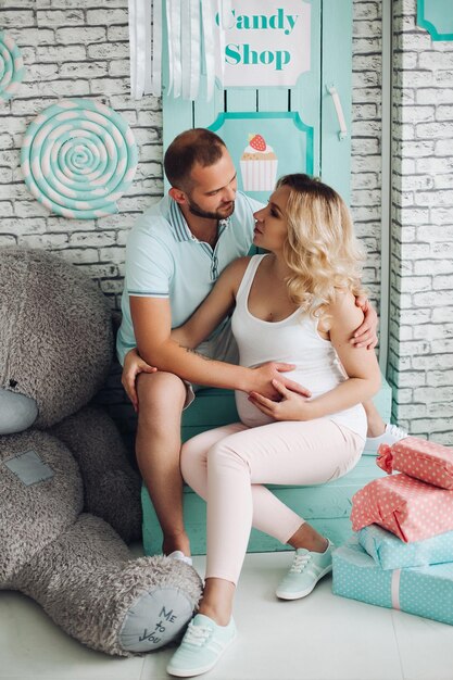 Couple de positivité de bel homme et jolie femme enceinte blonde assis dans les escaliers au studio embrassant l'estomac et gesticulant le cœur par les mains Jeune famille élégante à l'intérieur de la menthe douce parmi les jouets