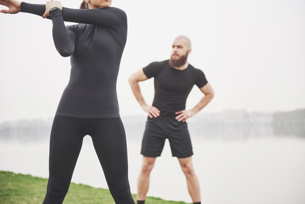 Couple de remise en forme qui s'étend à l'extérieur dans le parc près de l'eau. Jeune homme barbu et femme exerçant ensemble le matin