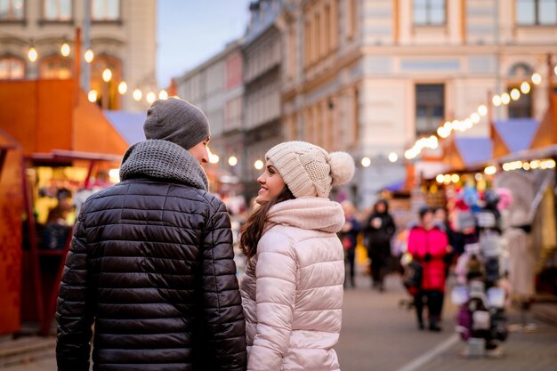 Couple romantique portant des vêtements d'hiver étreignant debout dans la rue du soir avec la foire de Noël