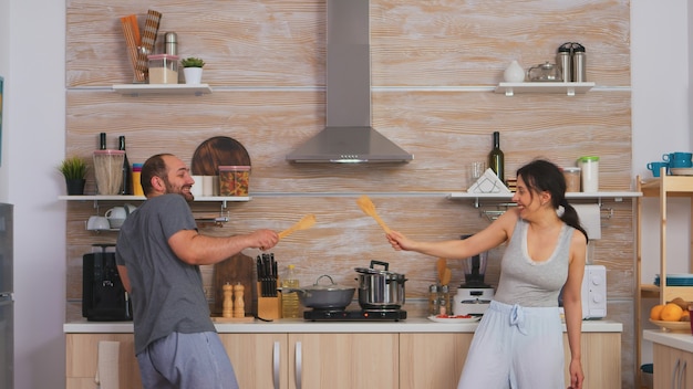 Couple s'amusant dans l'escrime de la cuisine avec de grosses cuillères pendant le petit-déjeuner en pyjama. Amants drôles joyeux et insouciants, cuillère en bois de combat, jeu de liaison, combat à l'épée, style de vie heureux