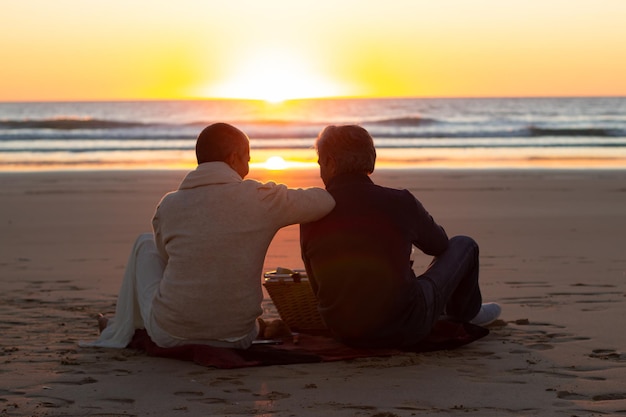 Couple senior romantique ayant pique-nique au coucher du soleil sur la plage. Homme et femme assis sur une couverture au bord de la mer et profitant d'une vue fantastique sur le paysage marin. Vue arrière. Romance, style de vie, concept de loisirs