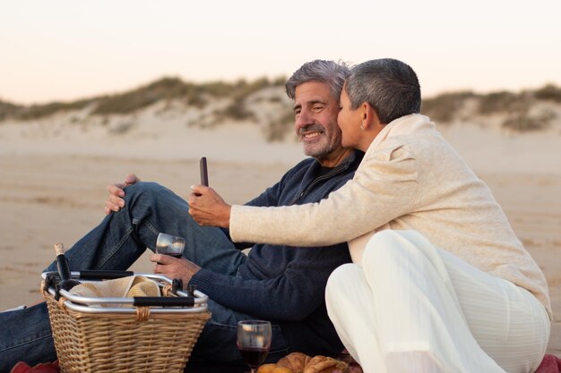 Photo gratuite couple senior romantique profitant d'un pique-nique au bord de la mer le soir, buvant du vin rouge et prenant un selfie. homme aux cheveux gris souriant tandis que dame avec smartphone embrassant sa joue. loisirs, concept romantique