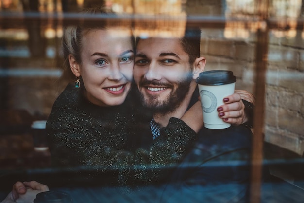 Un couple souriant et heureux est assis dans un café et regarde par la fenêtre tout en dégustant son café.