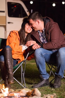 Couple souriant et profitant de la chaleur du feu de camp dans une froide nuit d'automne dans les montagnes avec un camping-car rétro en arrière-plan.