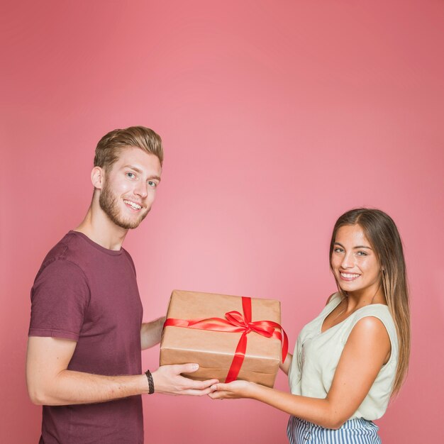 Couple souriant tenant la boîte cadeau avec ruban de satin rouge sur fond rose