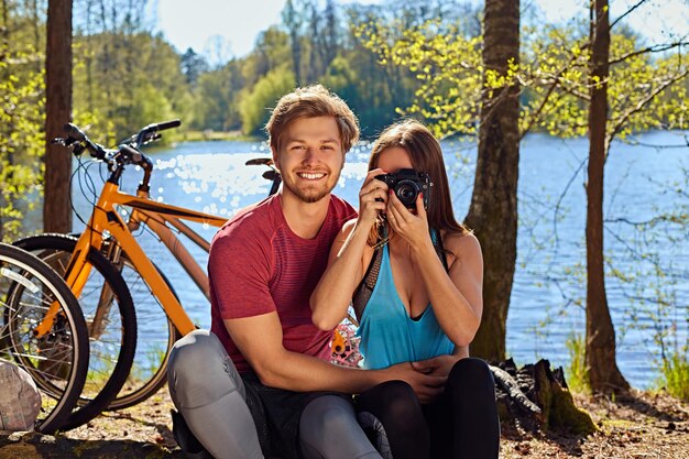 Couple sportif positif se relaxant près de la rivière après une balade à vélo. Femme prenant des photos sur un appareil photo numérique compact.