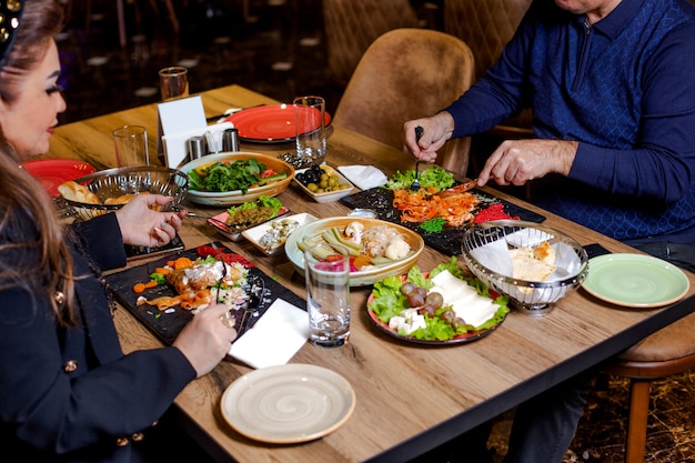 Couple en train de dîner avec des pâtes, salades, assiette de fromages, cornichons et olives