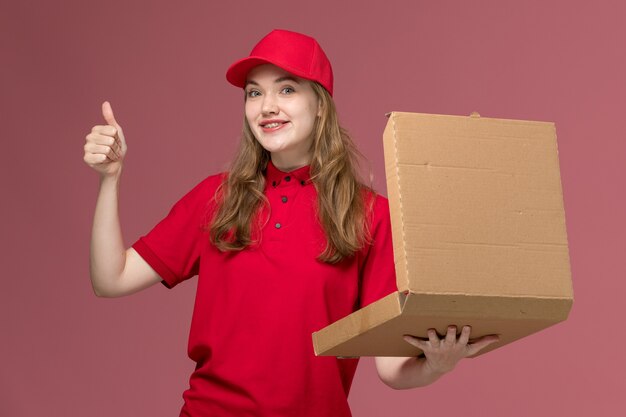 Courrier femelle en uniforme rouge tenant boîte de nourriture de livraison brune souriant sur rose clair, travail uniforme travailleur de service livraison fille