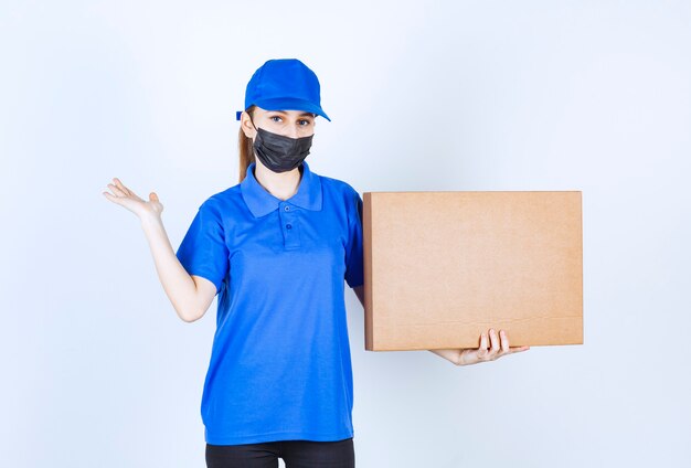 Courrier féminin en masque et uniforme bleu tenant un gros colis en carton et pointant vers quelqu'un