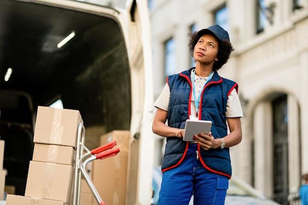 Courrier féminin noir utilisant une tablette numérique lors du déchargement des colis du camion de livraison dans la rue