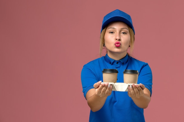 Courrier féminin en uniforme bleu posant et tenant des tasses de café s'embrasser sur rose, travail de livraison uniforme de service