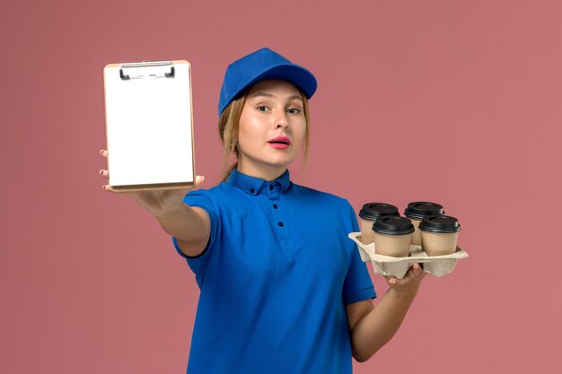 Courrier féminin en uniforme bleu tenant le bloc-notes avec des tasses de café marron sur rose clair, la livraison uniforme de service de travail