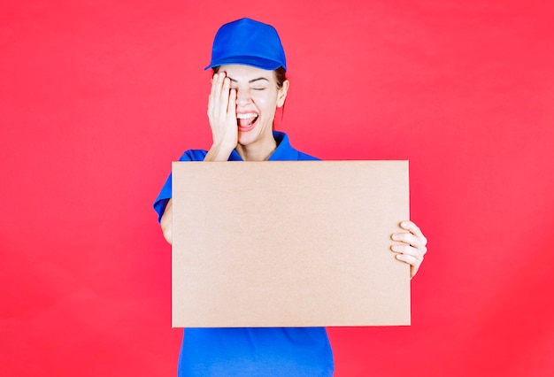 Courrier féminin en uniforme bleu tenant une boîte à pizza à emporter en carton et couvrant un œil avec la main.