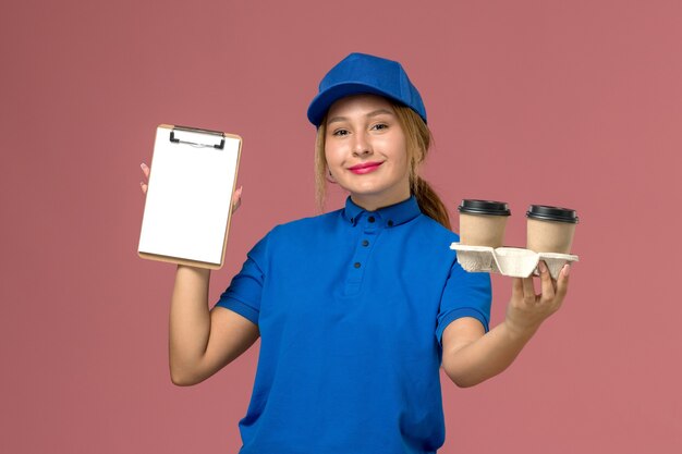 Courrier féminin en uniforme bleu tenant la livraison tasses de café et bloc-notes avec sourire sur rose, livraison uniforme de travailleur de service emploi