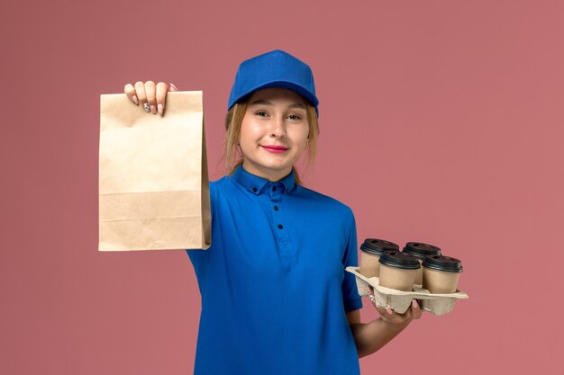 Courrier féminin en uniforme bleu tenant le paquet de nourriture et la livraison brune tasses de café sur rose, travailleur de travail de livraison uniforme de service